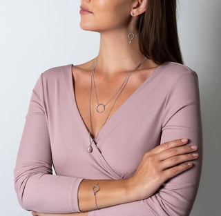 Woman wearing a pink top with jewelry including a necklace, earrings, and bracelet on a plain background.