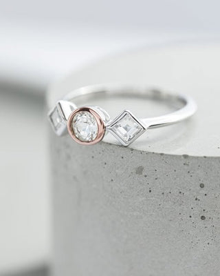 A silver ring with a round rose gold center stone and square baguette side stones, displayed on a textured gray cylindrical pedestal.