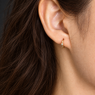 A close-up of a person's ear wearing a delicate gold huggie hoop earring with a small clear gemstone, set against dark brown hair and a neutral background.