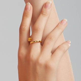 A close-up of a hand wearing a delicate yellow gold ring with a small, textured yellow gemstone, set against a soft gray background.