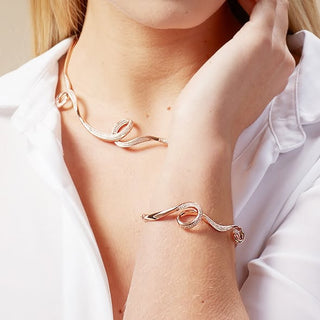 A close-up lifestyle shot of a model wearing a matching rose gold necklace and cuff bracelet with a twisted, snake-like design set with small clear crystals.