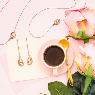 Rose gold jewelry set featuring a double-O pendant necklace, matching drop earrings, and a small heart-shaped pendant, styled in a flat-lay with a cup of coffee, pink roses, and a gold spoon on a light pink background.