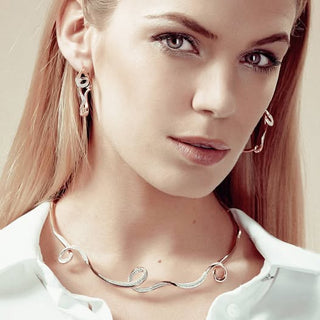 A close-up portrait of a woman wearing a rose gold wavy chain necklace and matching drop earrings, styled with a white collared shirt against a soft beige background.