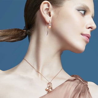 A close-up side profile of a woman wearing a rose gold necklace with a pearl pendant and matching drop earrings against a solid blue background.