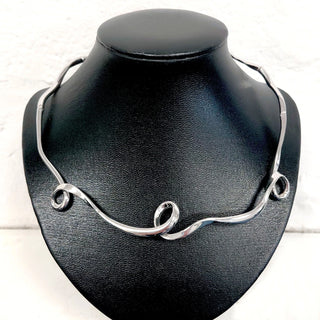 A polished silver wavy necklace displayed on a black jewelry bust against a white textured background.