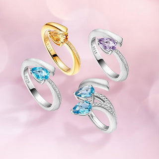 Four curved open-band rings arranged on a soft pink background. Two rings are silver-toned with heart-shaped blue gemstones and small clear stones along the band; one is silver-toned with a purple heart-shaped gemstone; one is yellow-toned with a pear-shaped yellow gemstone.