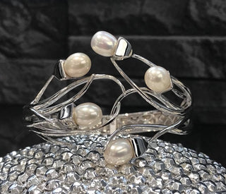 A silver-toned open cuff bracelet decorated with five white freshwater pearls, displayed on a sparkling silver crystal-covered surface against a dark textured background.