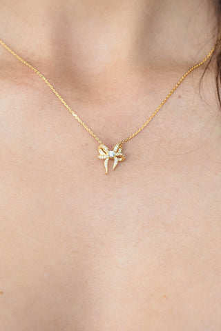 A close-up of a delicate gold chain necklace with a small bow-shaped pendant adorned with clear stones, worn on a model's neck.