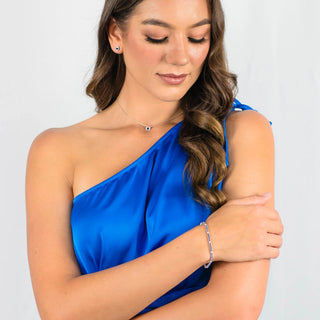 A woman wearing a bright blue one-shoulder satin dress, a delicate silver necklace, a silver stud earring, and a silver bracelet, shown against a plain light gray background.