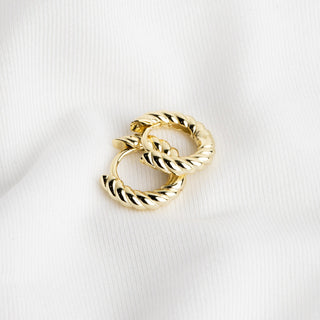 A pair of gold huggie hoop earrings with a twisted rope texture, displayed on a white textured fabric background.