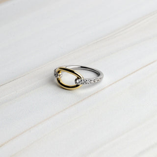 A two-tone ring featuring a polished gold and silver band, accented with small clear stones on the silver section, resting on a light wood surface.