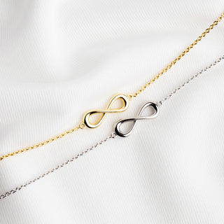 Two delicate infinity chain bracelets, one in yellow gold and one in silver, laid diagonally on a textured white fabric background.