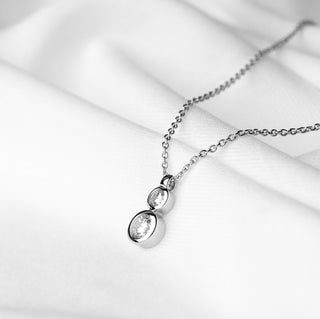 A silver necklace with a dual oval pendant resting on draped white fabric.