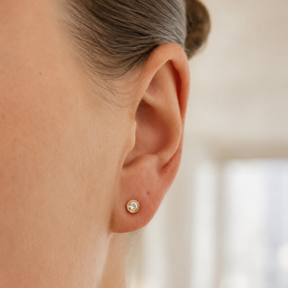 A close-up of a person's ear wearing a small gold stud earring with a clear gemstone, set against a softly blurred indoor background.