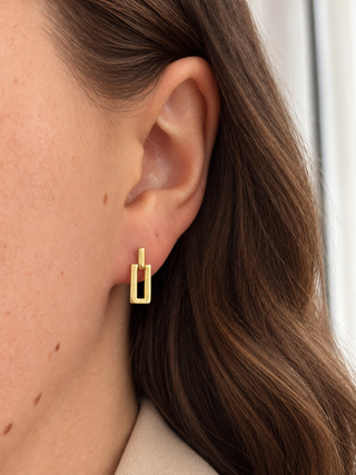 A close-up of a model's ear wearing a small gold geometric stud earring with a vertical rectangular frame design.