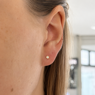 A close-up side profile of a woman's ear wearing a small round gold stud earring with a clear gemstone, set against a softly blurred indoor background.