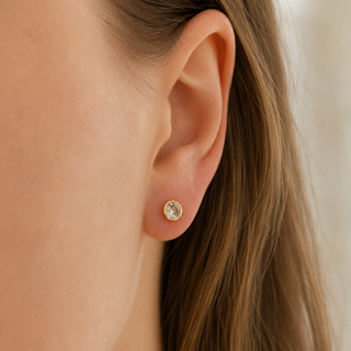 A close-up of a model's ear wearing a small gold stud earring with a clear round gemstone.