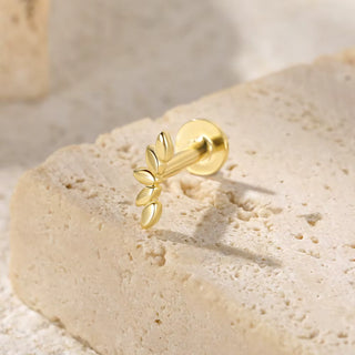 A gold leaf-shaped labret stud earring resting on a textured beige stone block.