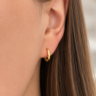 A close-up of a model's ear wearing a small, polished gold huggie hoop earring.