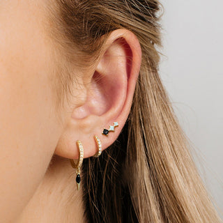 Close-up of an ear wearing gold hoop earrings with black stones on a neutral background