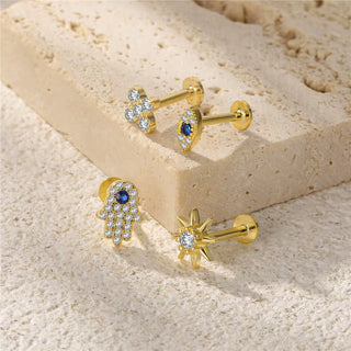 Three gold-plated stud earrings displayed on a textured beige stone slab and sandy surface. The earrings include a hamsa hand with a blue gemstone and clear crystals, a small blue oval gemstone surrounded by clear crystals, and a starburst design with a clear central gemstone.