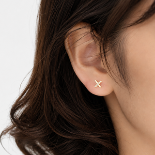 A close-up of a woman's ear wearing a small gold stud earring shaped like an 'X', with her dark brown hair against a clean white background.