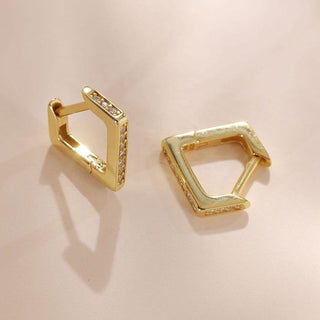 A pair of gold geometric hoop earrings with small clear stones set along the front edges, displayed on a light beige background.
