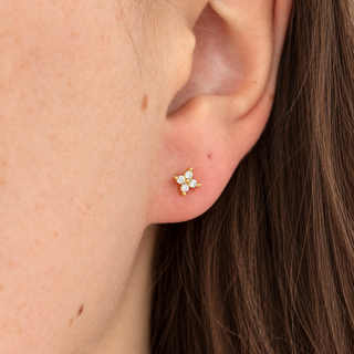 A close-up of a person's ear wearing a small gold stud earring with three clear gemstones, set against skin and brown hair.