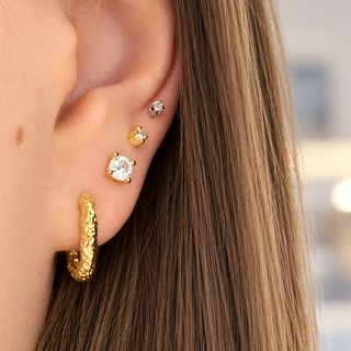 A close-up of a person's ear showcasing four gold and silver stud earrings, including a textured gold huggie hoop, a gold stud with a clear gemstone, a small gold stud, and a silver stud with a clear gemstone.