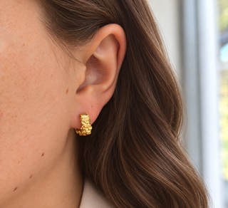 A close-up of a woman's ear wearing a small textured gold huggie hoop earring, with her brown hair and skin visible against a softly blurred light background.