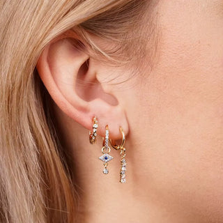 A close-up of a model's ear wearing three gold huggie hoop earrings, including one with a dangling eye motif set with a blue stone and clear crystals.