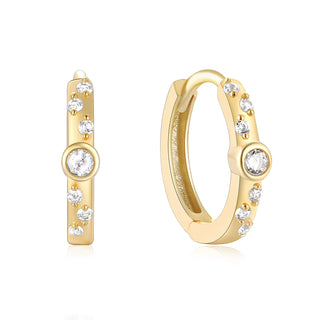 A pair of gold huggie hoop earrings with clear round-cut gemstones, shown on a clean white background. One earring is shown from a front-facing perspective, and the other is angled to show the inner band and side details.