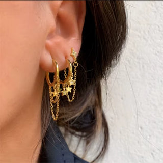 A close-up of a person's ear wearing multiple gold hoop and chain earrings with dangling star charms, against a light textured wall.