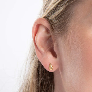 A close-up of a person's ear wearing a small gold teardrop stud earring against a clean white background.