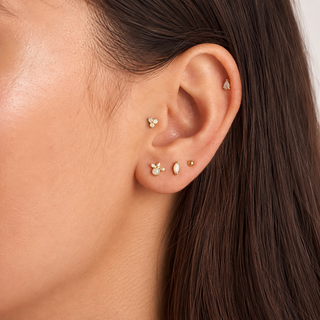 Close-up of a person's ear showcasing multiple delicate gold ear piercings, including a small stud, a tiny dome, a butterfly cluster, and a teardrop pearl stud, against dark brown hair.