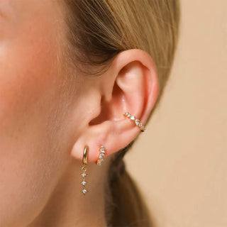 Close-up of a person's ear wearing multiple gold ear piercings: a small hoop drop earring with clear stones, a curved ear cuff with clear stones, and a small vertical cluster stud.
