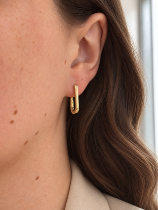 A close-up of a woman's ear wearing a small gold huggie hoop earring, with her brown hair and beige top visible in the background.