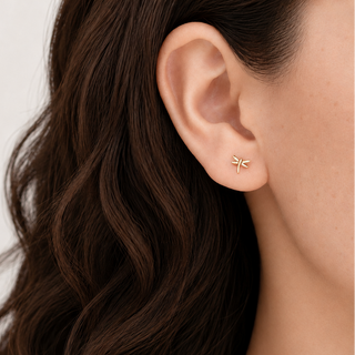 A close-up of a woman's ear wearing a small gold dragonfly stud earring, with her dark brown wavy hair and skin visible against a clean light background.
