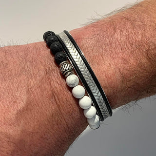 A close-up of a man's wrist wearing three stacked bracelets: a black lava stone bead bracelet, a black and silver chevron-patterned metal bracelet, and a black bracelet with white round beads and a silver spacer bead.