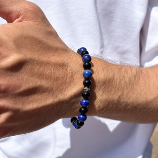 A close-up of a beaded stretch bracelet worn on a person's wrist. The bracelet features alternating polished royal blue and matte black round beads, with a small silver-toned spacer bead near the center.