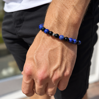 A close-up of a man's wrist wearing a beaded bracelet made of alternating dark blue lapis lazuli and black beads, paired with a white t-shirt and black shorts.