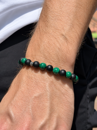 A close-up of a beaded stretch bracelet worn on a person's wrist. The bracelet features alternating matte black beads and polished green beads, including a green-and-black banded stone bead.