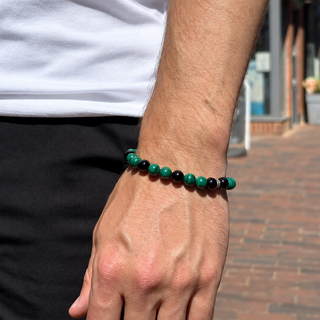 A close-up of a person's wrist wearing a beaded bracelet made of alternating dark green and black beads, set against a blurred outdoor street background.