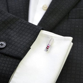 A close-up of a white dress shirt cuff with a silver-toned tie bar featuring a small red and white striped accent, styled on a dark patterned suit jacket.
