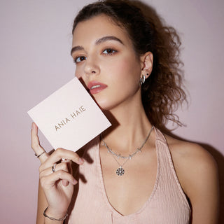 Woman holding a 'ANIA HAIE' product with jewelry on a plain background