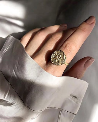 Hand wearing a gold ring with intricate design, wearing a light-colored shirt.
