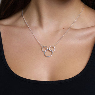 A close-up of a person wearing a delicate silver necklace with three interlocking circular rings at the center of the chest, paired with a black scoop-neck top.