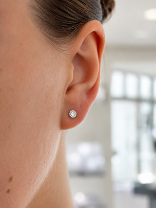 A close-up side profile of a person's ear wearing a small round silver stud earring with a clear gemstone, set against a softly blurred indoor background.