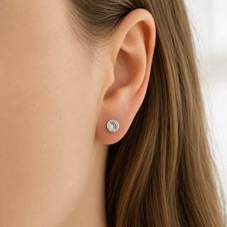 A close-up of a person's ear wearing a small round stud earring with a clear gemstone in a silver-toned setting.