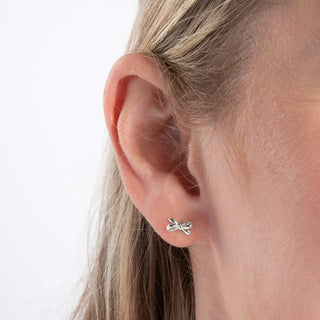 A close-up of a person's ear wearing a small silver bow-shaped stud earring against a plain white background.